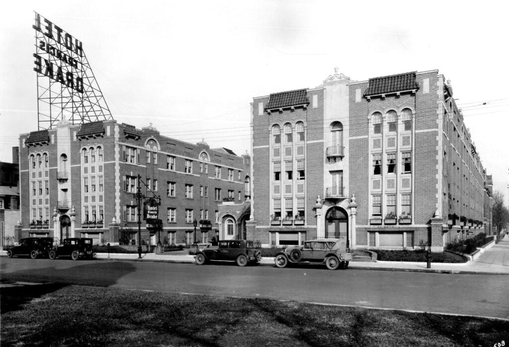 Black and white photo of two brick hotels with vintage cars parked in front and a large “Hotel Drake” sign on the roof.