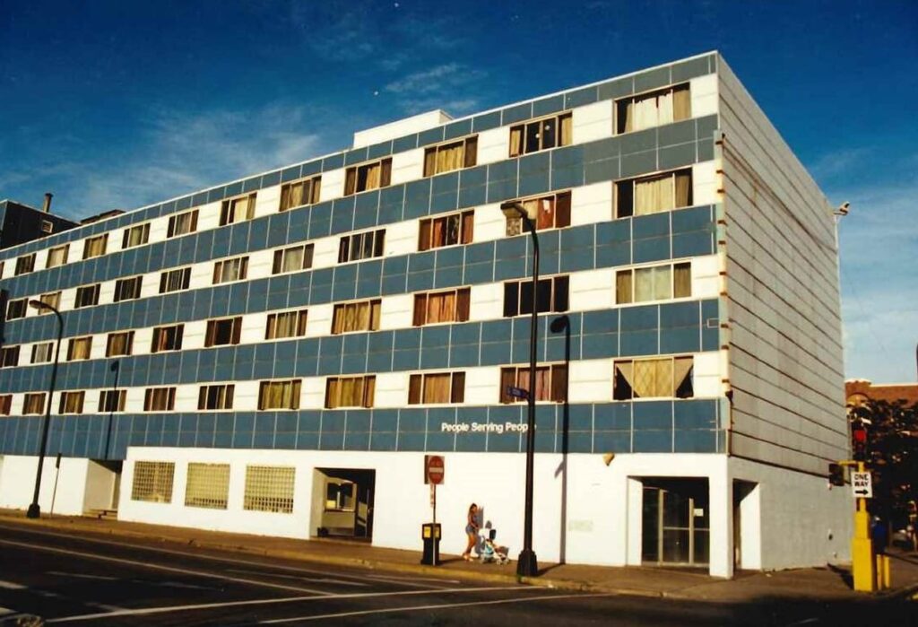 A blue and white four-story building with many windows, two people walking on the sidewalk in front.