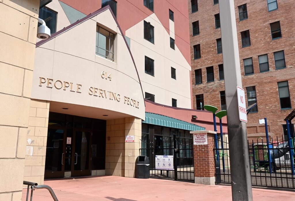 Entrance to a building with "People Serving People" above the door, surrounded by fencing and tall brick buildings.