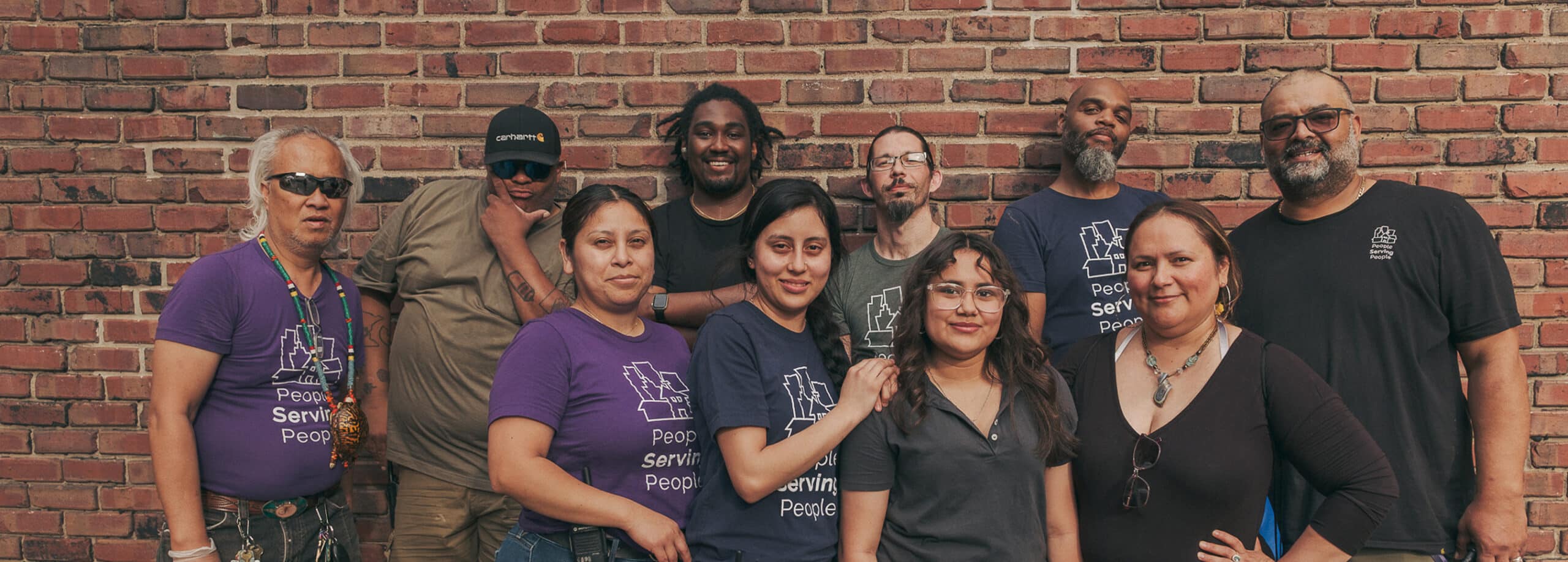 A diverse group of ten people pose together, smiling, in front of a brick wall. Some wear "People Serving People" shirts.