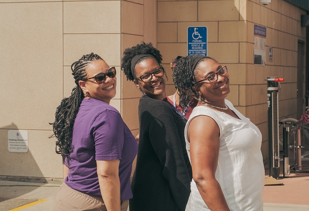Three women smiling and posing together outdoors near a building and a blue accessible parking sign.