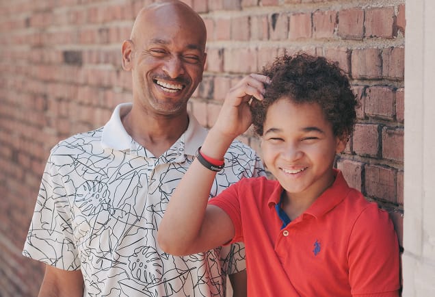 Two people, an adult and a child, smiling and standing together against a brick wall on a sunny day.