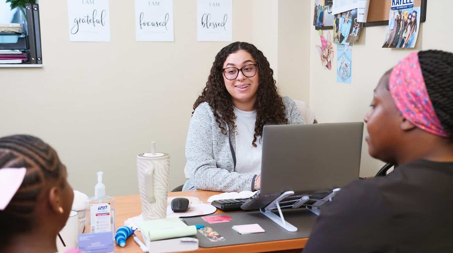 A woman at a desk talks to two people; motivational posters and photos are on the wall behind her.