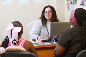 A woman at a desk smiles while talking to a young girl and another woman sitting across from her.