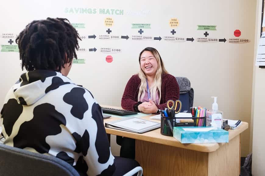 Two people sit across a desk, smiling and talking, in an office with a savings match chart on the wall behind them.