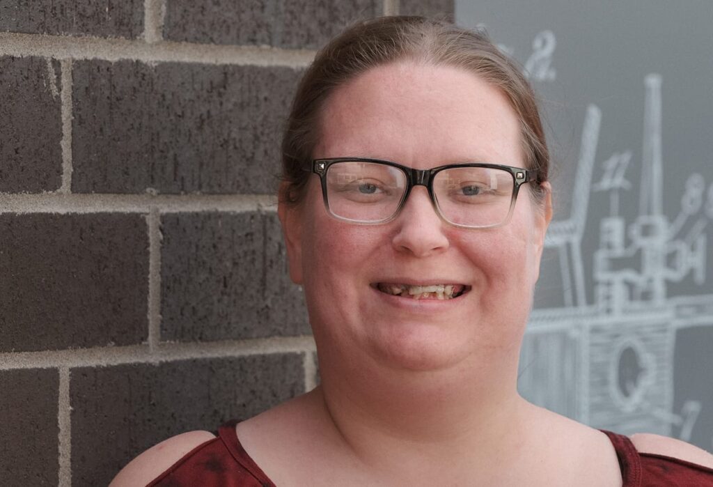 A person with glasses smiles in front of a brick wall and a gray background with white line drawings.