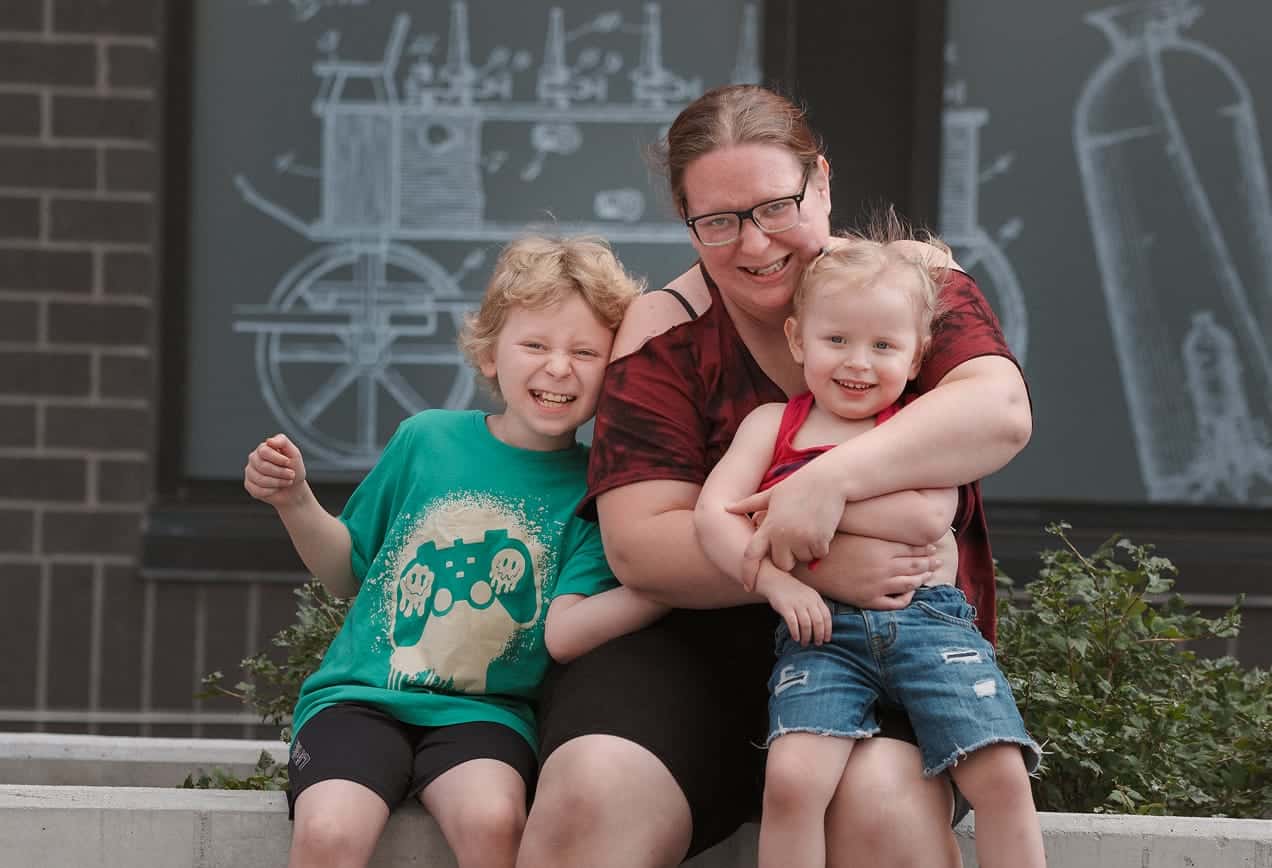 A smiling adult hugs two happy children while sitting outside in front of a wall with science-themed drawings.