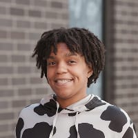 Smiling person wearing a black and white patterned hoodie, standing in front of a brick wall.