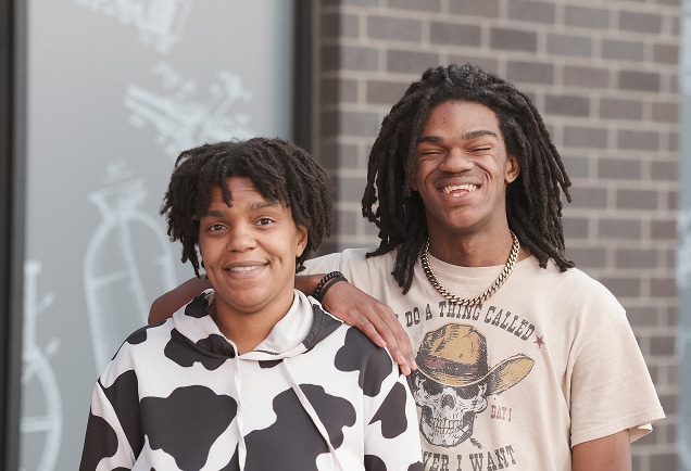 Two people with dreadlocks smiling, one wearing a cow-print hoodie, the other a graphic t-shirt, standing by a brick wall.