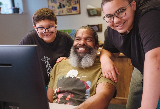A smiling man with a beard and two boys wearing glasses gather around a computer, looking happy and engaged together.