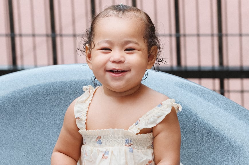 Smiling baby with curly hair in a light dress, sitting on a blue chair outdoors in front of a black metal fence.