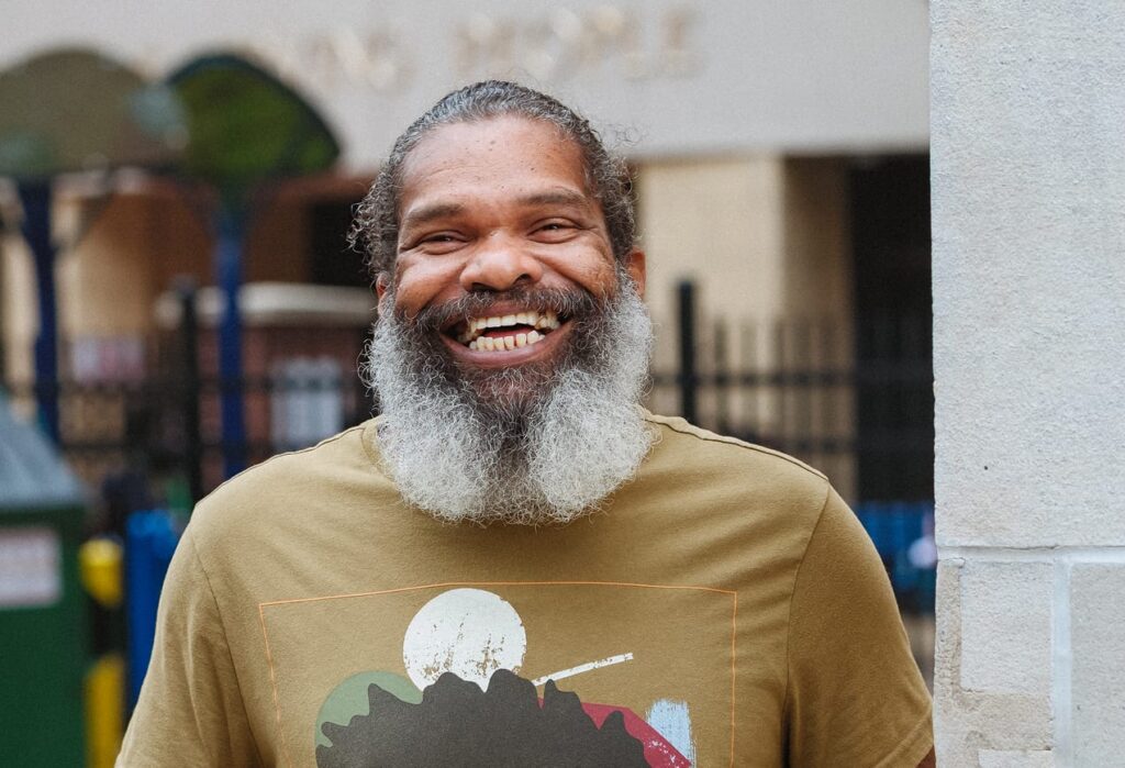 Smiling man with a gray beard wearing a graphic t-shirt stands outdoors in front of a building and a black fence.