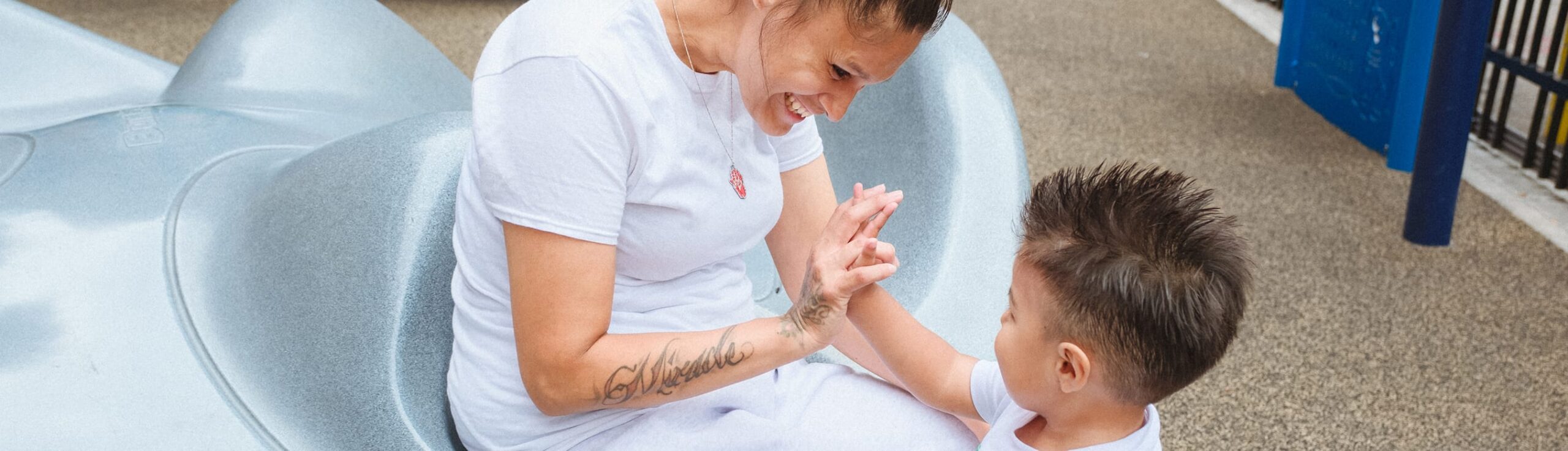 Woman and young boy smiling and playing together on playground equipment outdoors.