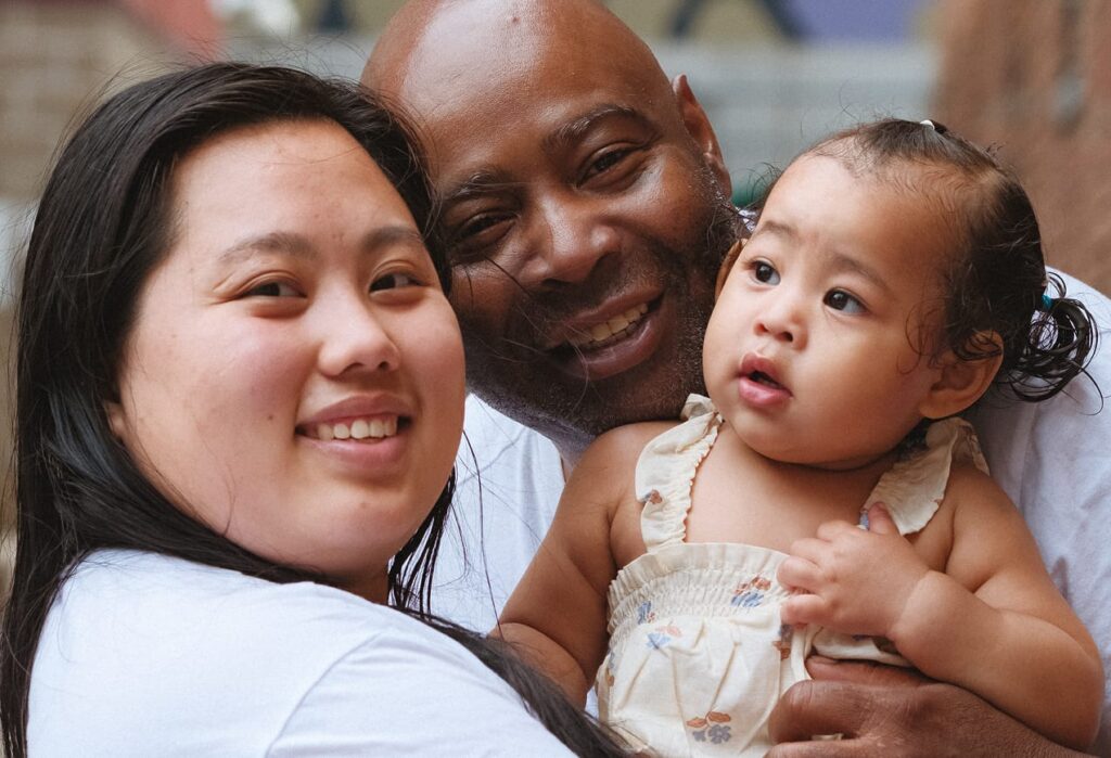 A woman, man, and baby smile and pose closely together outdoors, showing warmth and affection.