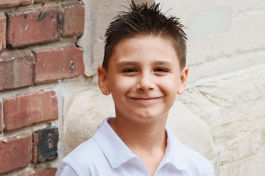 Smiling young boy with spiky hair and a white shirt stands in front of a brick and stone wall.