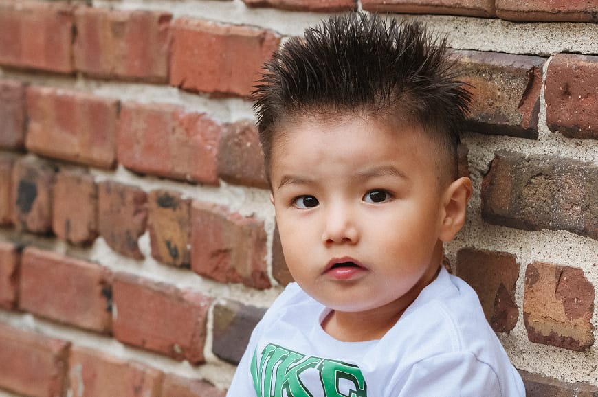 Toddler with spiky hair and a white shirt sits in front of a brick wall, looking slightly to the side.