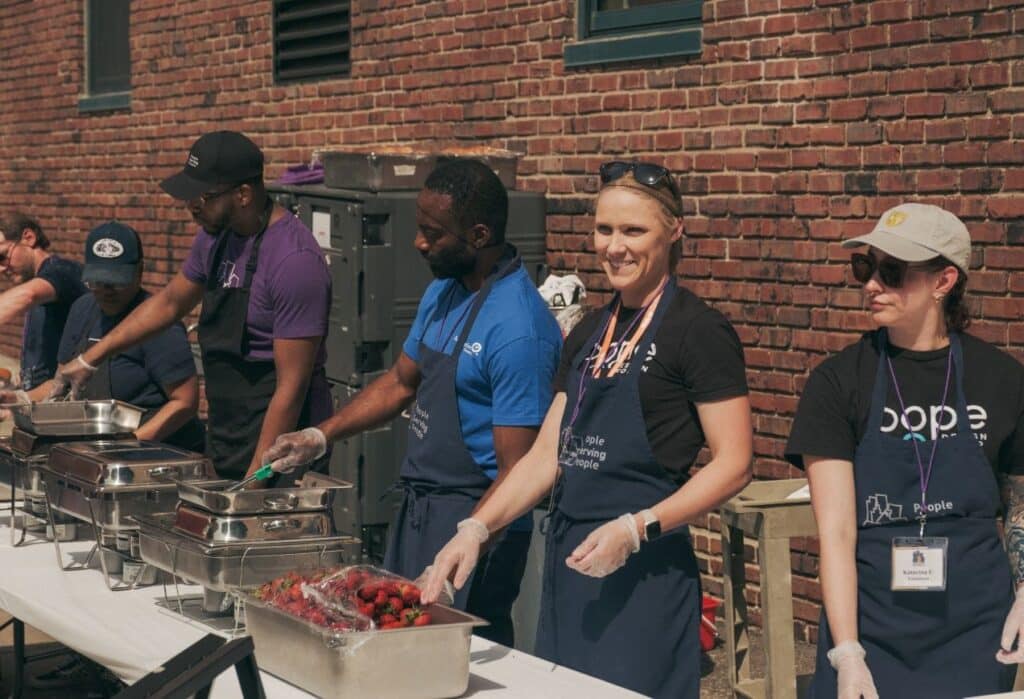 Five people wearing gloves and aprons serve food outdoors at a long table against a brick wall.
