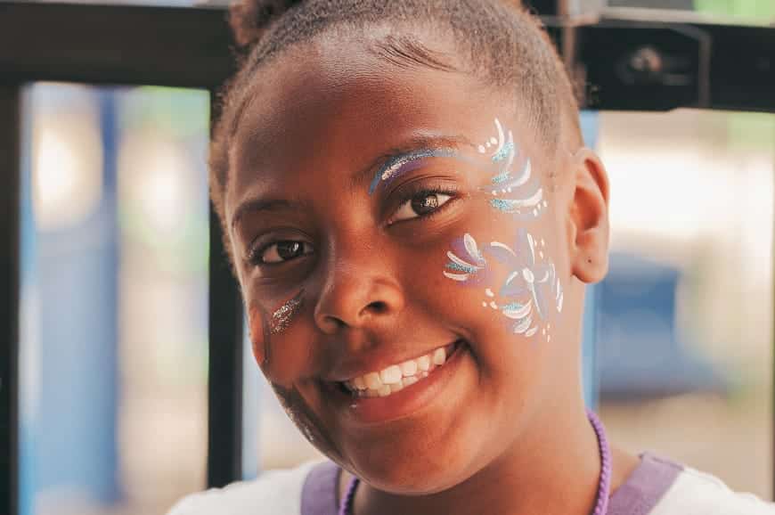 Smiling girl with colorful face paint designs on her cheeks and forehead, standing outdoors.