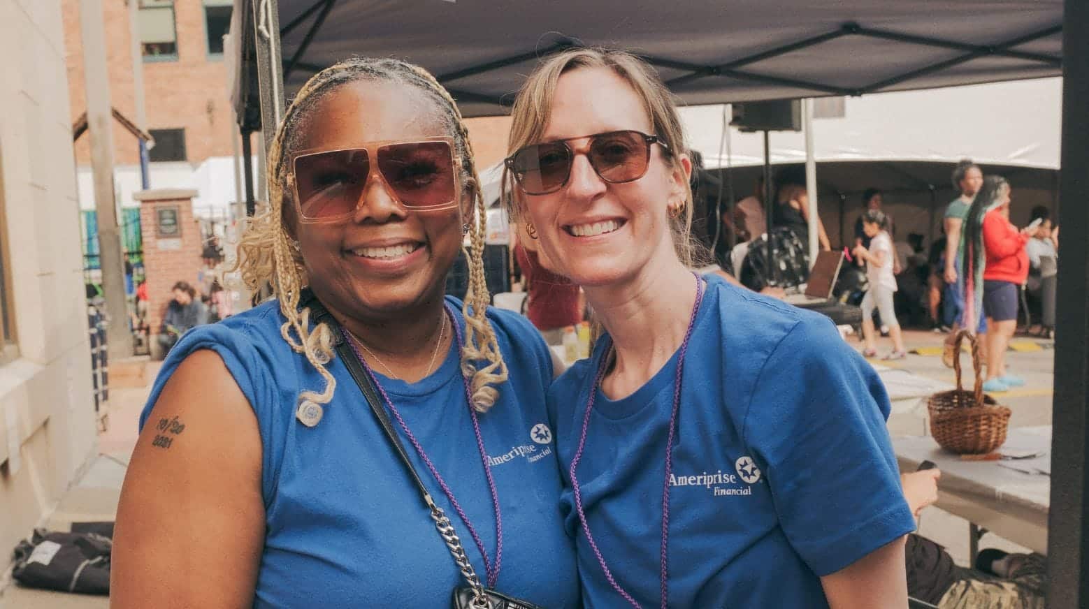 Two smiling women in blue Ameriprise Financial shirts pose together outdoors at an event, wearing sunglasses.