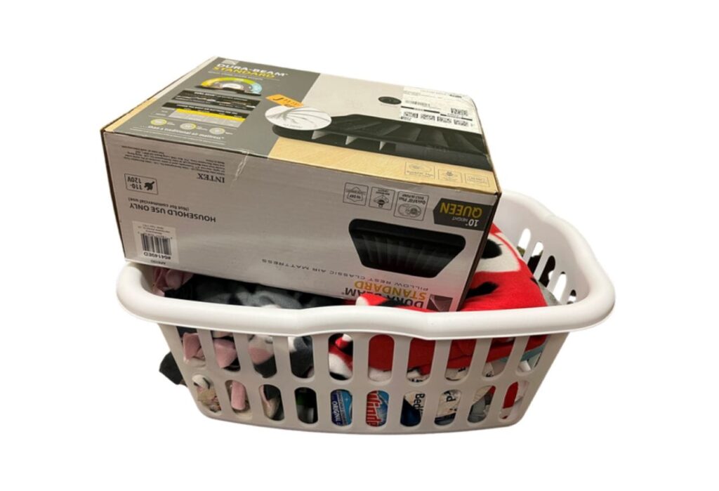 A white laundry basket filled with clothes and a boxed air mattress placed on top.