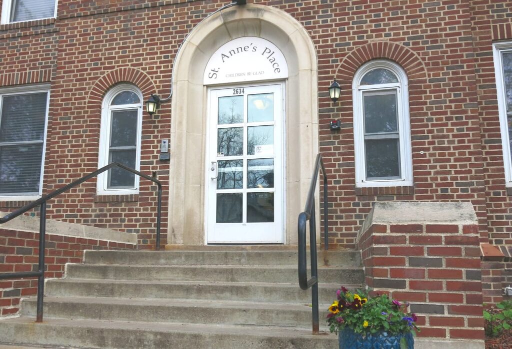 Entrance to a brick building with steps, a white door, and a sign reading “St. Anne’s Place.”.