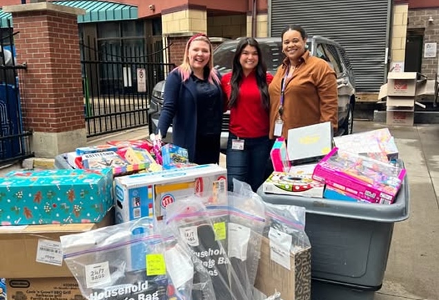 Three women smile behind bins and boxes filled with toys and gifts outside a building on a cloudy day.