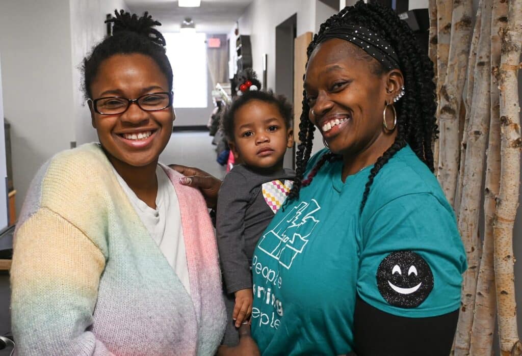 Two smiling women and a young girl pose together indoors, with one woman holding the child.