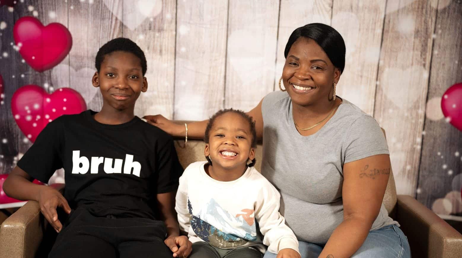 Smiling woman and two children pose together in front of a heart-themed backdrop.