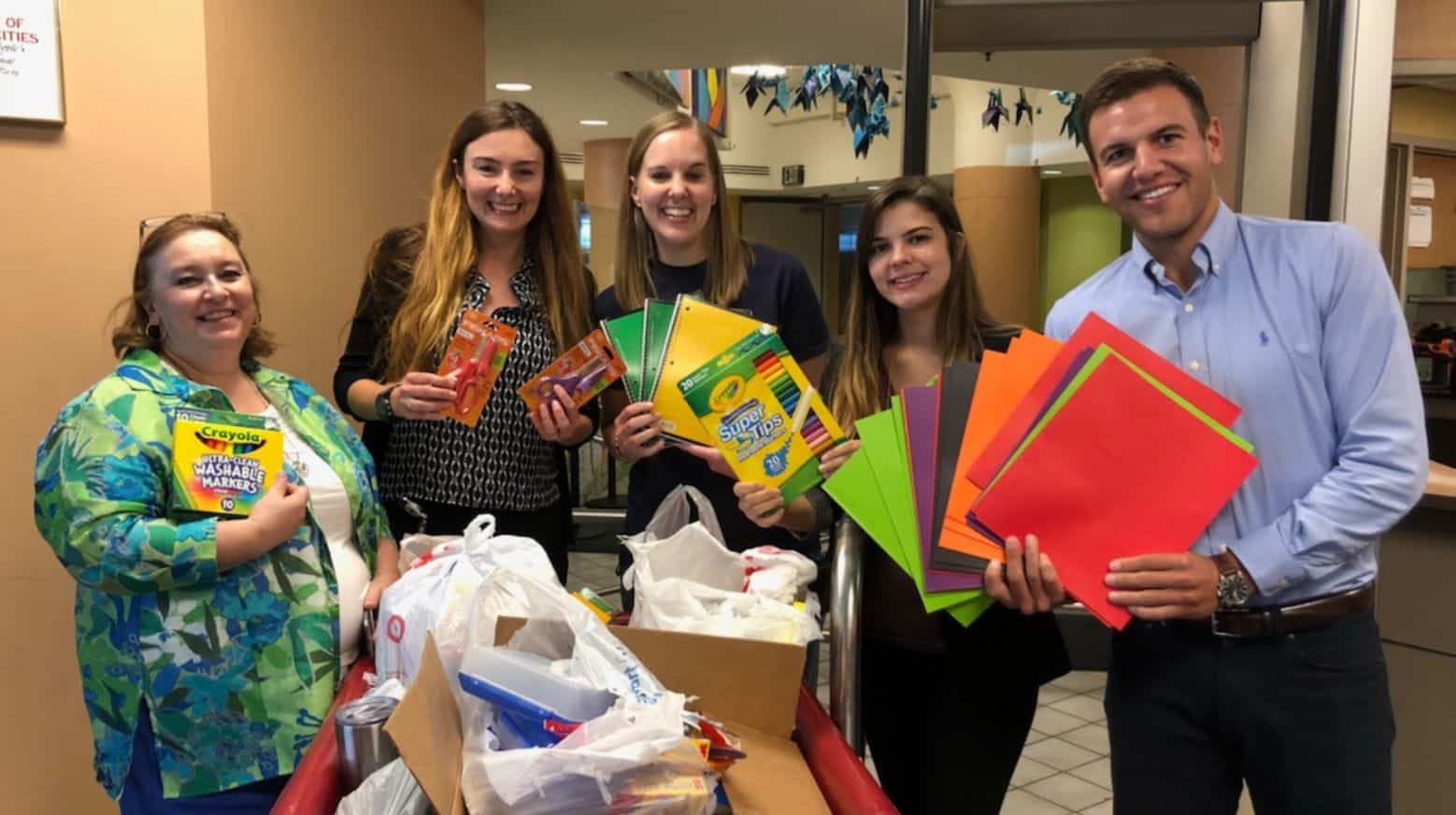 Five smiling people hold school supplies and colorful paper next to a cart filled with bags in a hallway.
