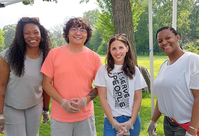 Four people smiling outdoors, wearing gloves, standing together under trees on a sunny day.