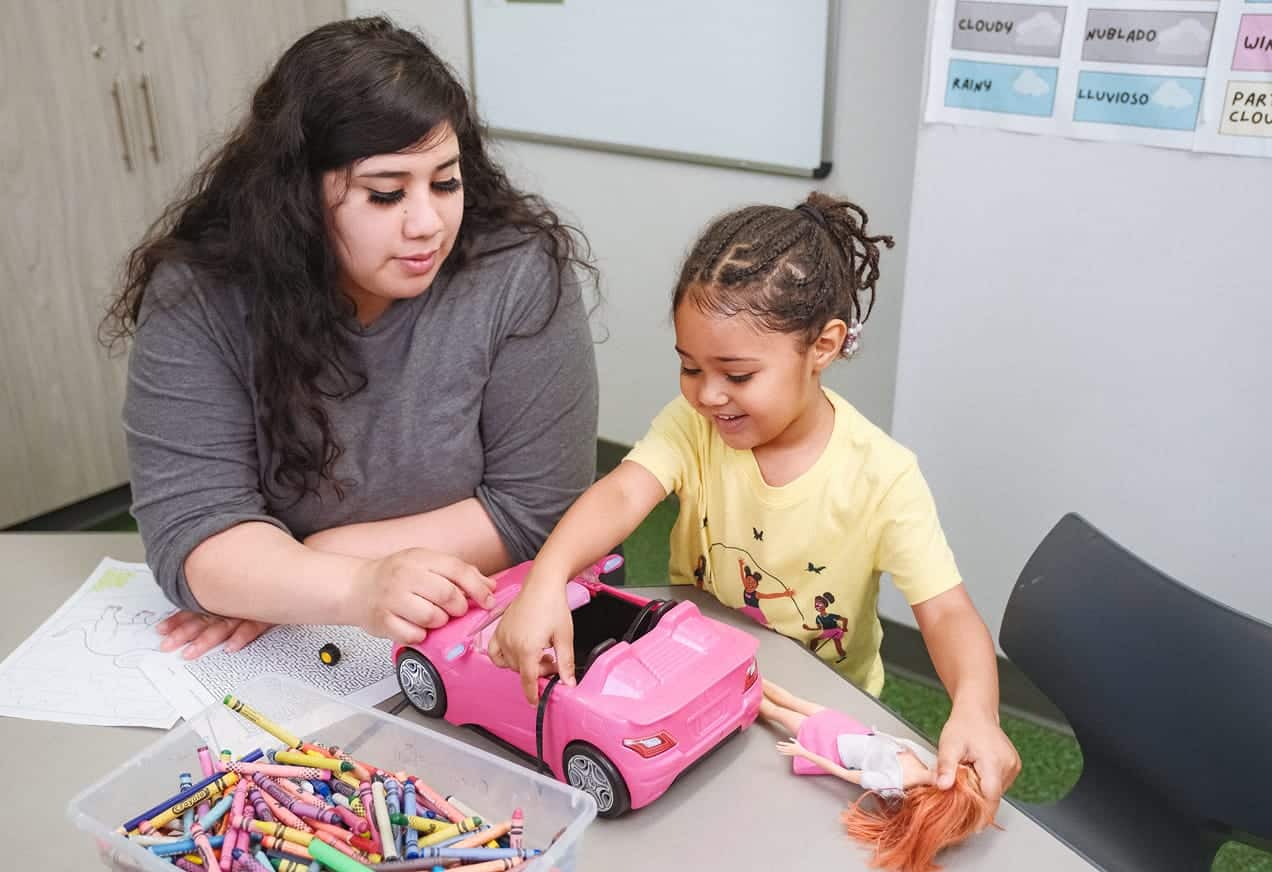 An adult and a child play with a toy car and doll at a table covered with crayons and coloring pages.