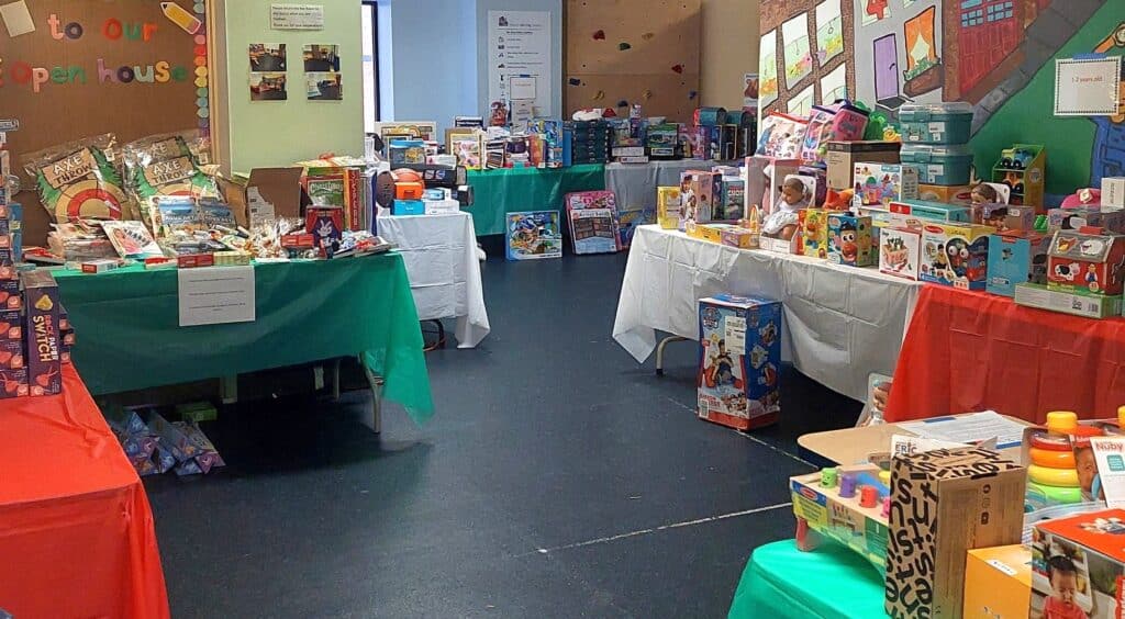 Rectangular tables with white, green, and red table coverings are lined against the walls of a children's rec room, which is colorfully painted. On these tables lies piles of toys for all ages.