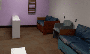 A room with old blue chairs and a blue couch surrounded by light purple and white walls.