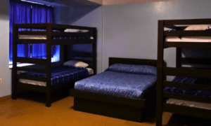 A brightly lit room with 2 bunk beds and a full size bed with blue comforter and dark wood frames.
