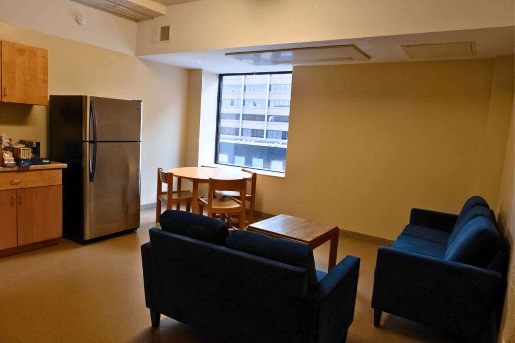 A beige room contains a wooden table and chairs by a window. Two blue couches surround a small coffee table. To the left, there is a large grey fridge and kitchen cabinets.