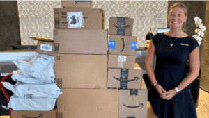 A woman in a black dress stands in a fancy lobby next to a rolling cart filled with Amazon boxes containing shelter donations.