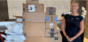 A woman in a black dress stands in a fancy lobby next to a rolling cart filled with Amazon boxes containing shelter donations.