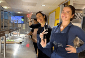 Two women in blue aprons smile as they stand in line behind a serving station at a cafeteria.