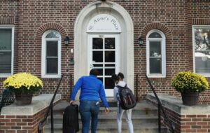 A woman and her daughter carry luggage up the front steps of a brown brick building, with "St. Anne's Place" painted over the white doorway.