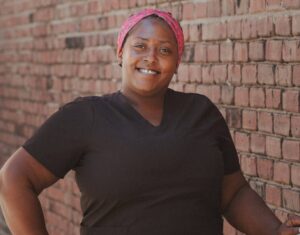 A woman wearing a black t-shirt and black leggings, with her hair wrapped behind a pink headband, smiles in front of red brick wall.