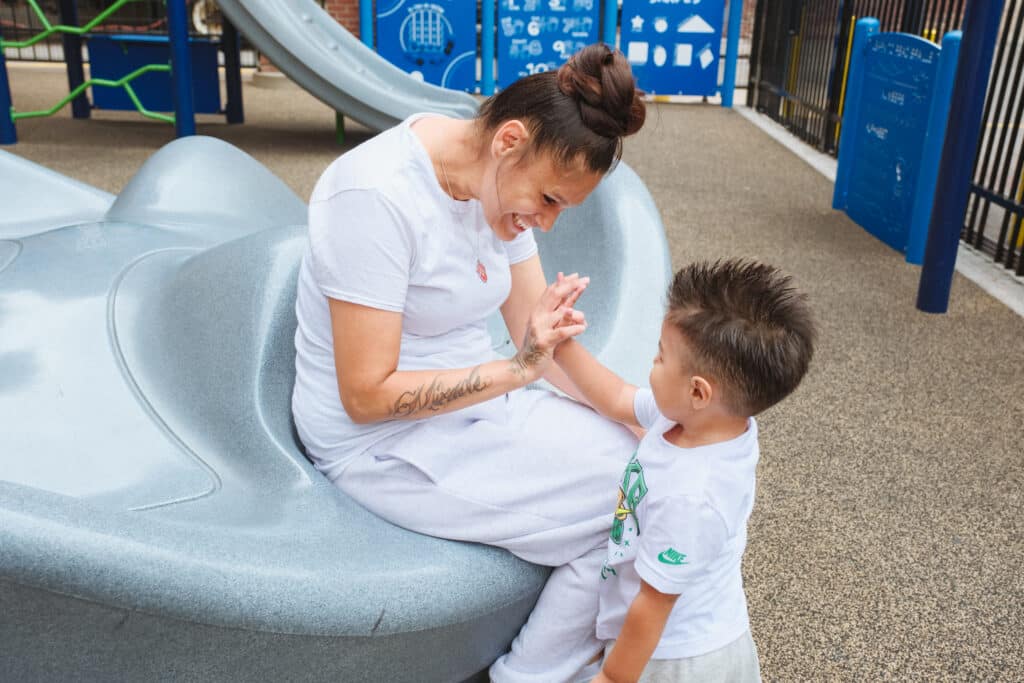 A mom and son are on a playground, both wearing white. The mom, with her dark hair in a ponytail, smiles and gives her young son a high-five.