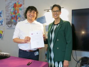 A man in a white button down shirt and a woman in a green blazer stand next to each other, holding a signed piece of paper.