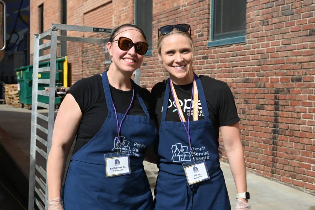 Two young women with ponytails and sunglasses smile outside and don blue aprons, ready to serve food.