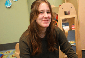 A young woman with long, wavy brown hair and a green long-sleeve sweater smiles with a classroom in the background.
